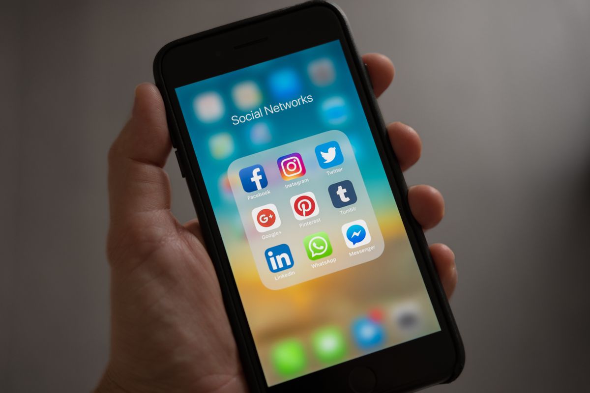 A hand holds a smartphone displaying a folder of social media app icons, illustrating the platforms where comment spam appears