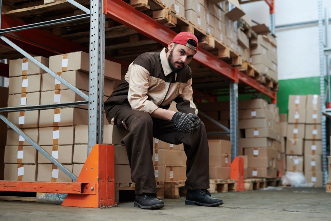 Warehouse worker sitting on a wooden pallet surrounded by stacked cardboard shipping boxes