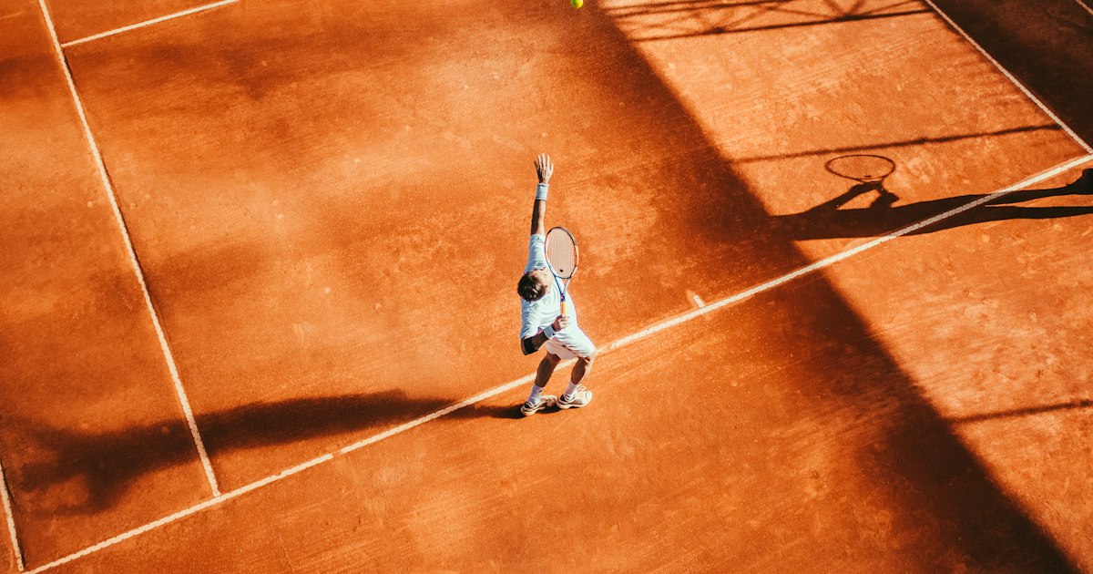 Tennis player mid-serve on an outdoor hard court with racket raised overhead