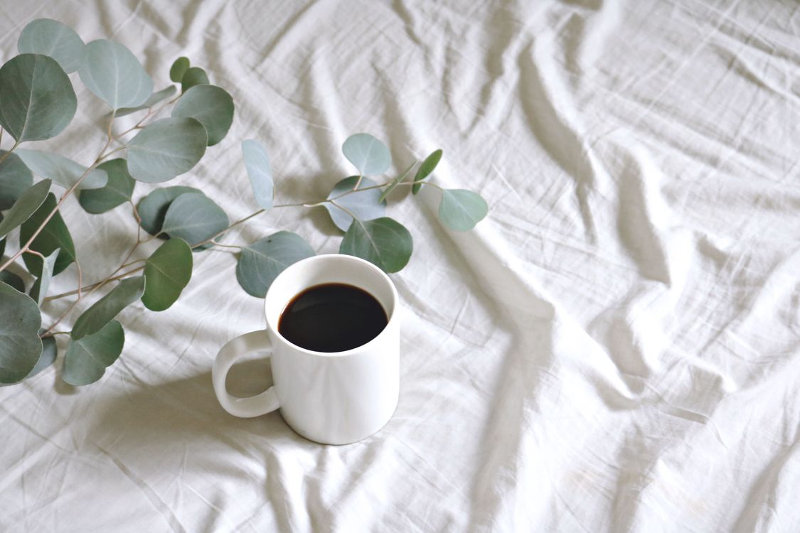 White ceramic coffee mug with black coffee beside eucalyptus leaves on linen