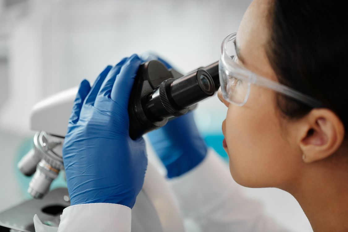Pesquisadora com óculos de proteção examinando amostra ao microscópio de laboratório