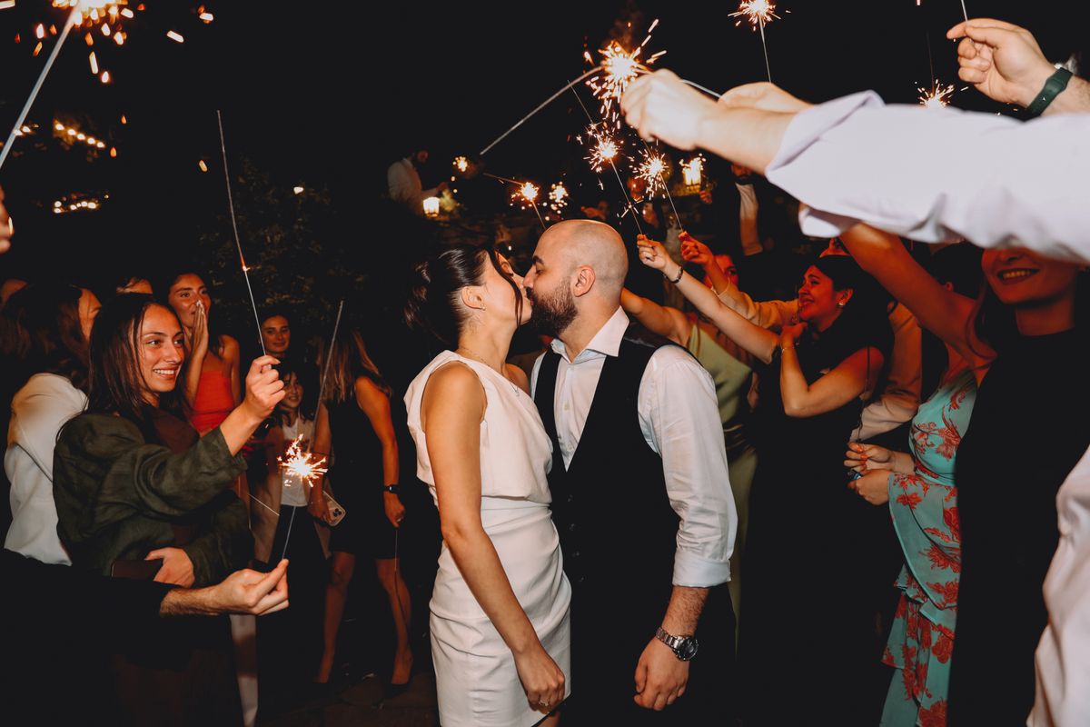 Wedding guests holding sparklers surround newlyweds dancing at a nighttime reception