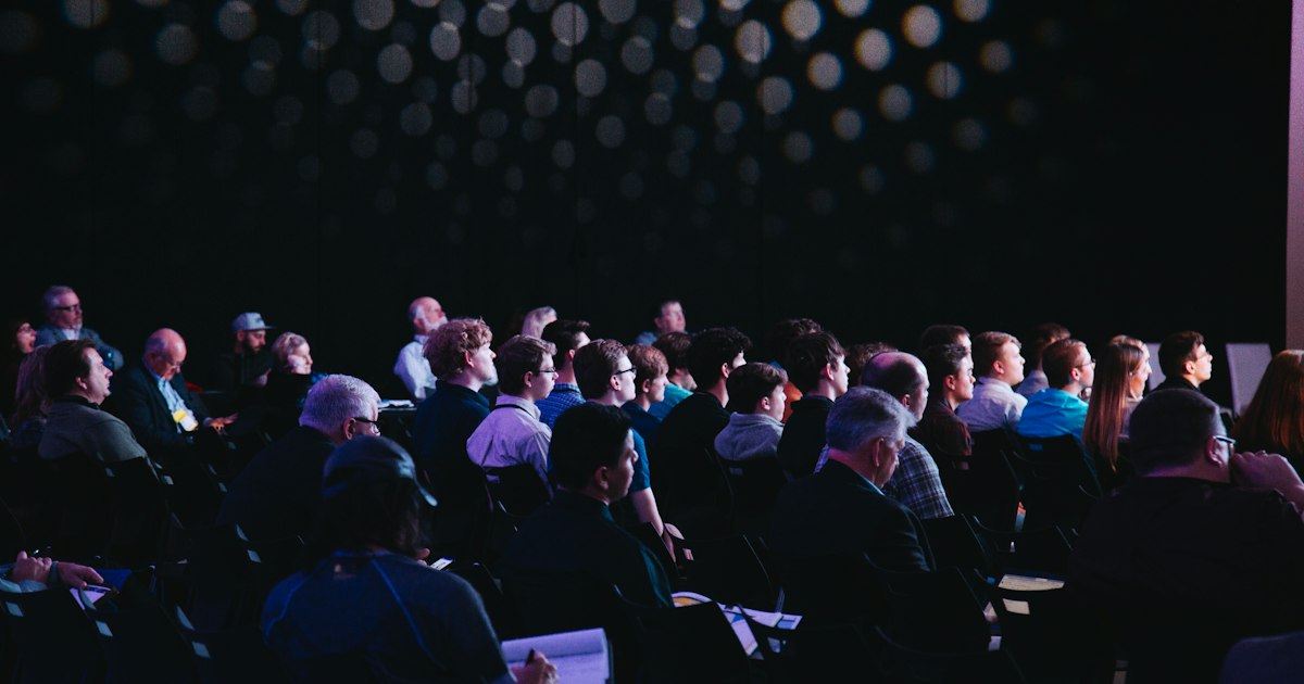 Professional conference hall with rows of attendees facing a brightly lit presentation stage